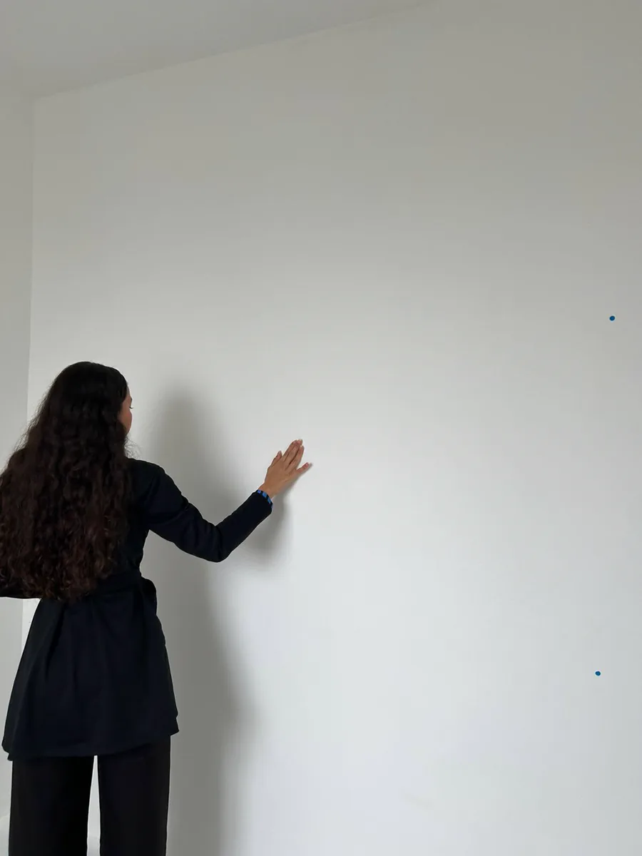 Bianca com a mão apoiada em uma parede branca marcada com pontos de laser, verificando planicidade.