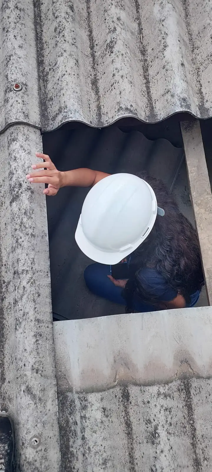 Vista de cima de Bianca, de capacete, inspecionando o interior de uma cobertura por uma abertura entre telhas.