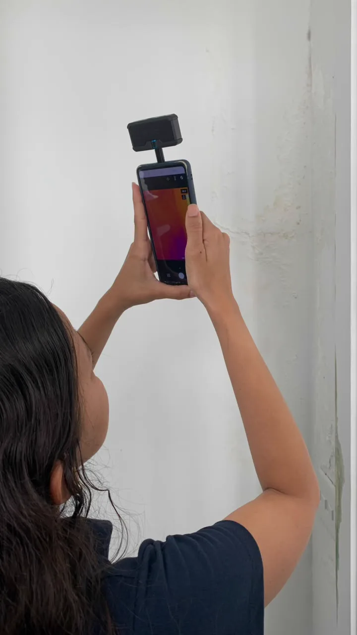 Câmera termográfica acoplada a um celular sendo apontada para uma parede com mancha de infiltração.